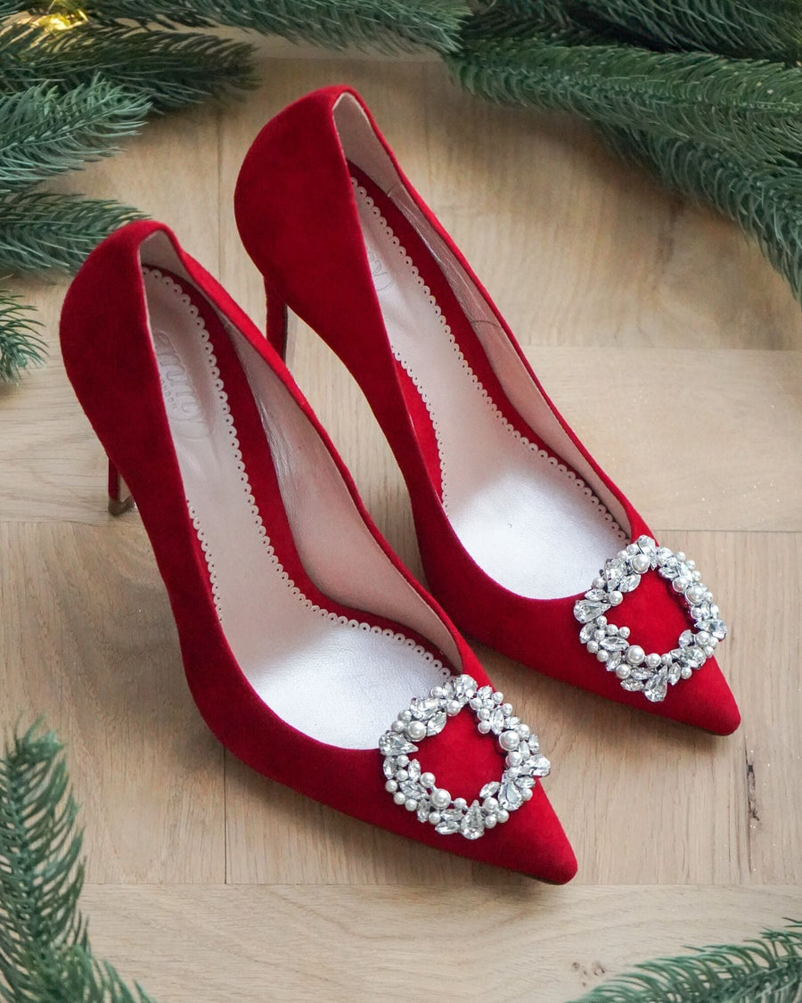 Red suede high heels with a pointed toe and embellished buckle detail on the front, displayed on a wooden surface surrounded by green foliage. The image captures the shoes from a top-down angle, showcasing their elegant design. image