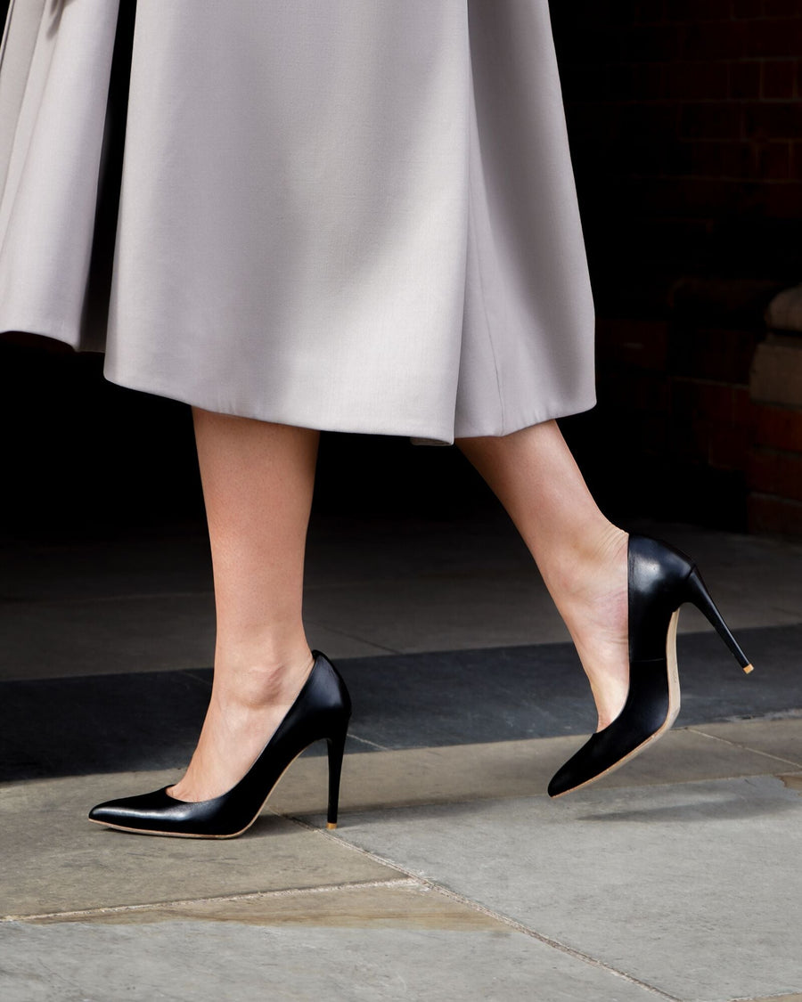 Black leather high heels are worn by a model walking on a stone surface, with a light gray skirt flowing around her. The image captures her from the ankle down, showcasing the sleek design of the shoes against the backdrop of a brick wall. image