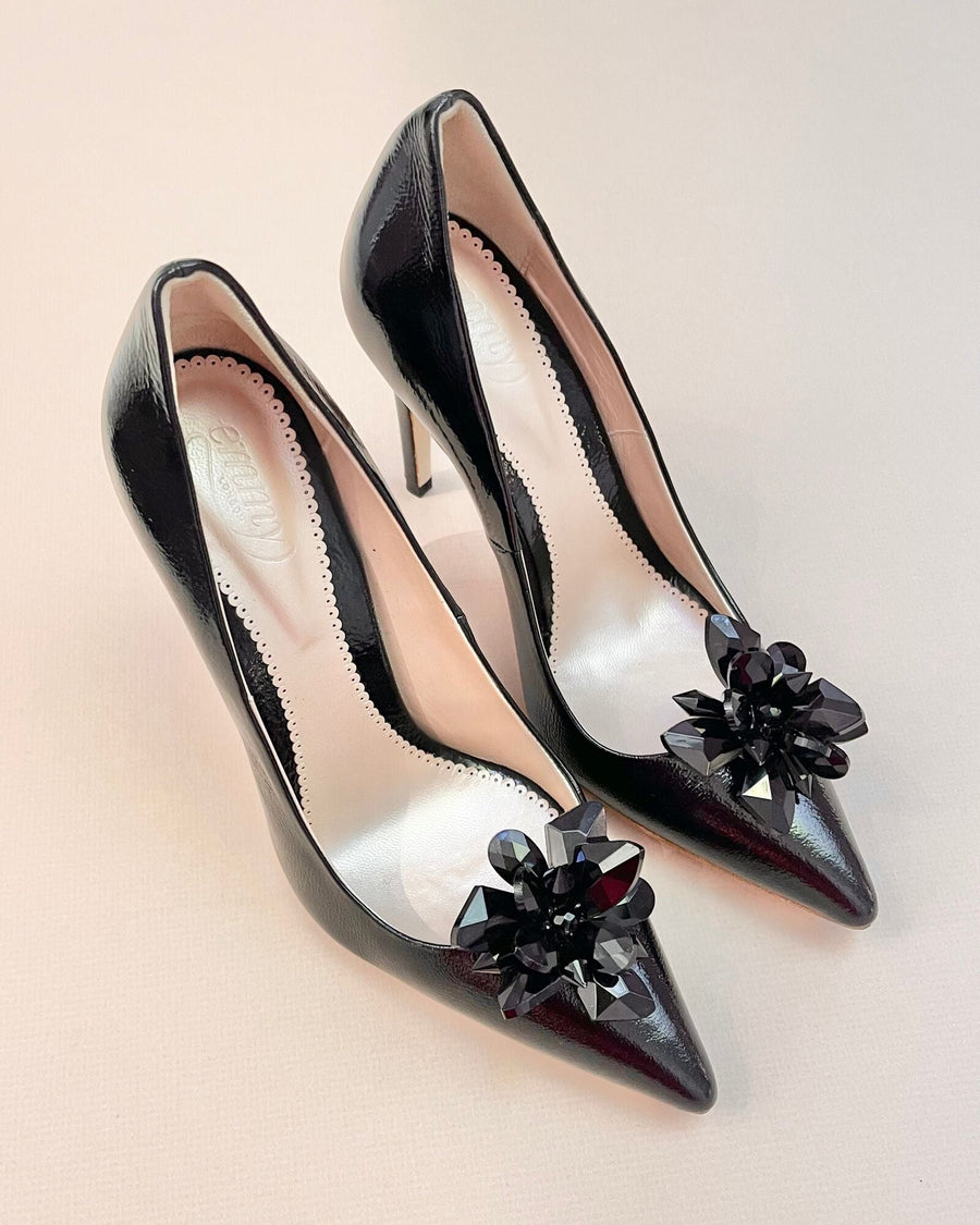 Black patent leather high heels featuring a pointed toe and decorative floral embellishments on the front. The shoes are displayed against a neutral background, showcasing their sleek design and elegant silhouette. image