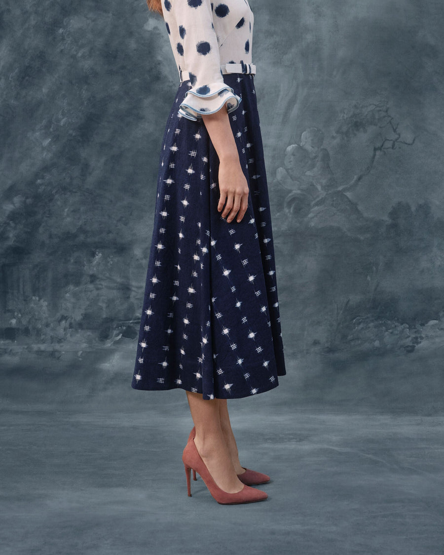 Elegant pink suede heels with a pointed toe, featuring a sleek stiletto heel. The model is standing in profile against a textured gray background, showcasing the shoes alongside a flowing navy skirt adorned with white polka dots and a matching blouse. The angle highlights the height of the heels, complementing the overall outfit. image