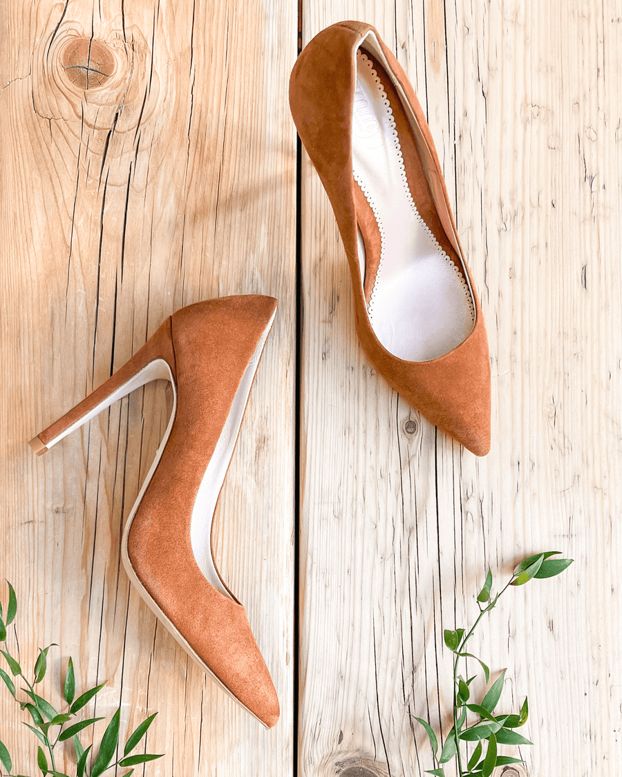 Brown suede high heels are displayed on a wooden surface, with a natural background featuring greenery. The shoes are pointed-toe and have a sleek design, showcasing their elegant silhouette from a top-down angle. image