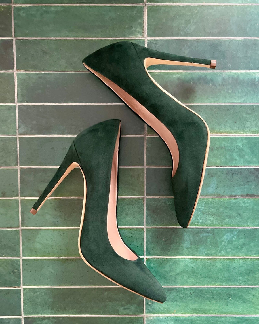 Green suede high heels are displayed on a textured green tile background. The shoes feature a pointed toe and a sleek silhouette, showcasing their elegant design. image