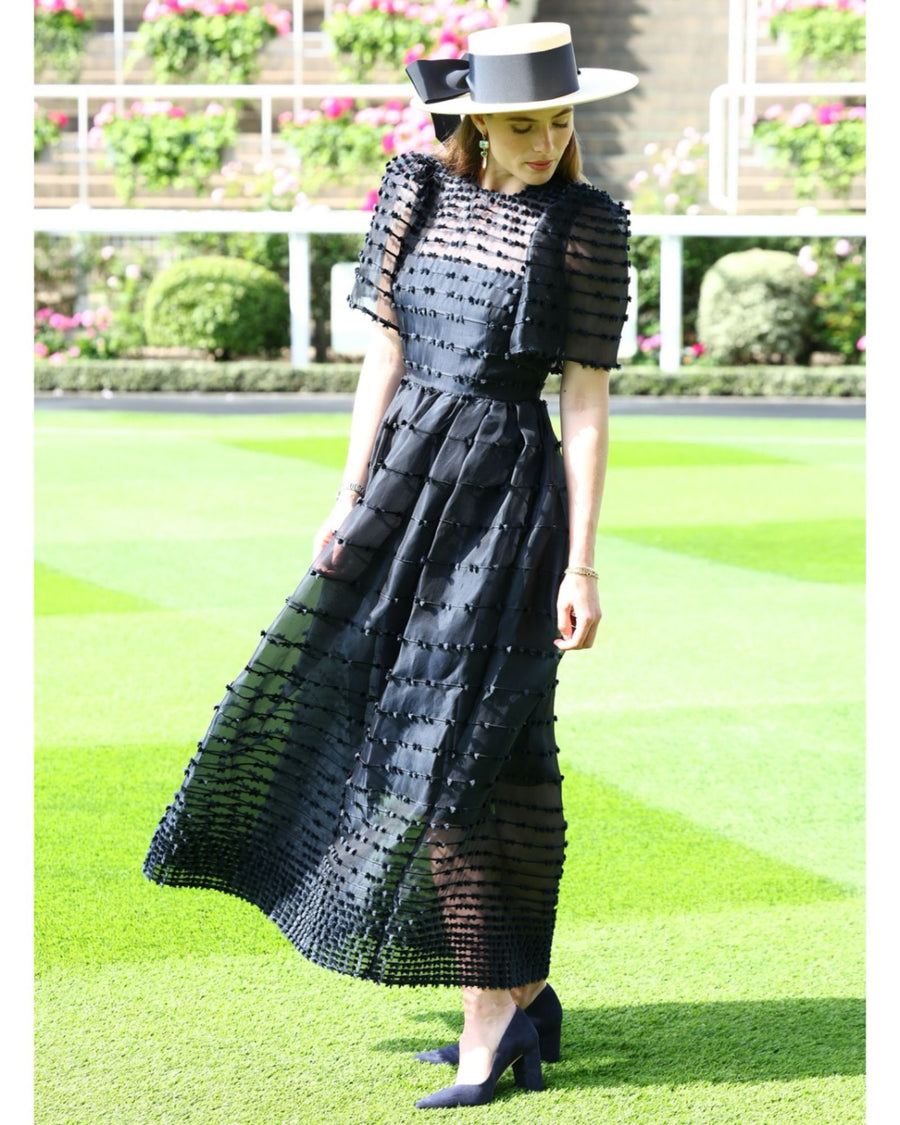 A woman is wearing a black, floor-length dress with a textured design, standing on a lush green lawn. She has a wide-brimmed hat adorned with a bow. The image captures her in profile, showcasing the elegant silhouette of the dress and the stylish heels she is wearing. The background features vibrant flower arrangements and manicured greenery, adding to the sophisticated atmosphere. The heels are made of leather. image