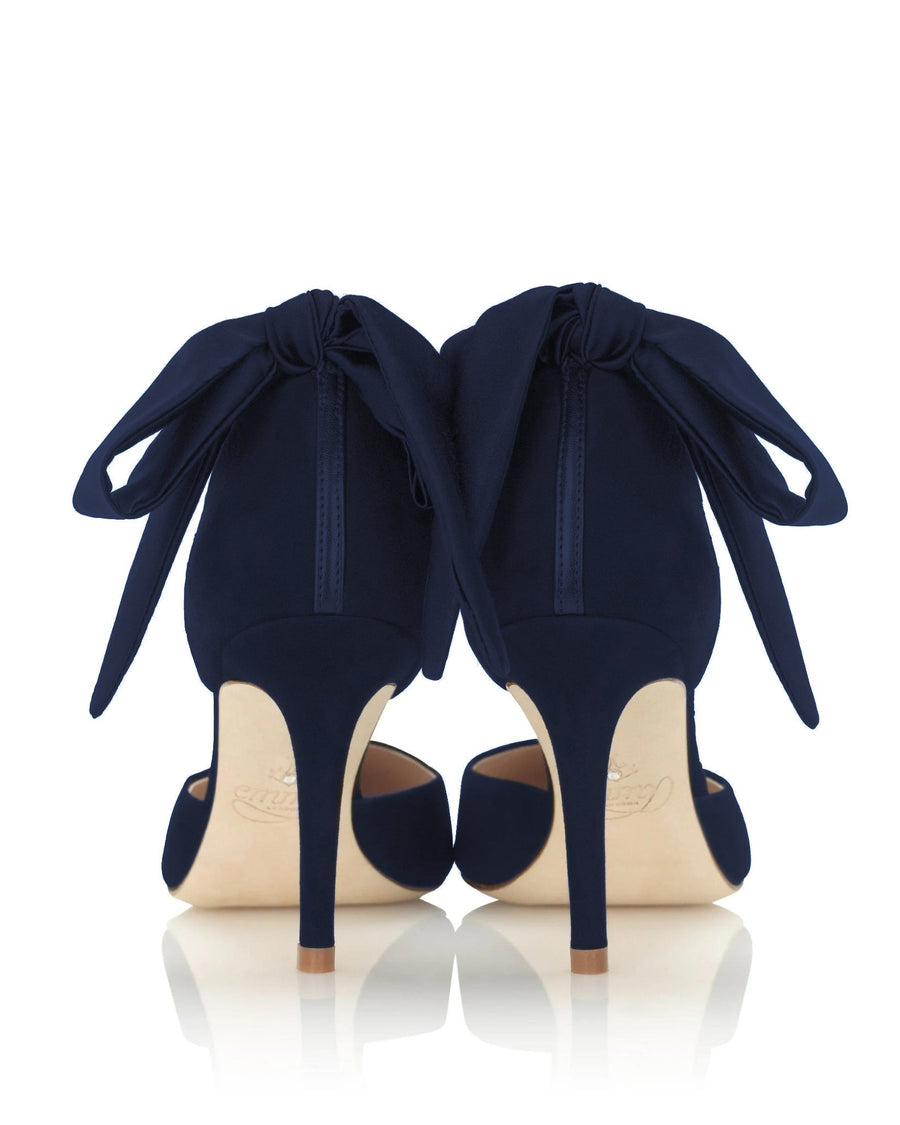 Navy blue heels with a sleek design featuring a large bow at the back. The shoes have a slender stiletto heel and are made of suede. The image is taken from a low angle, showcasing the elegant silhouette against a clean, white background. image