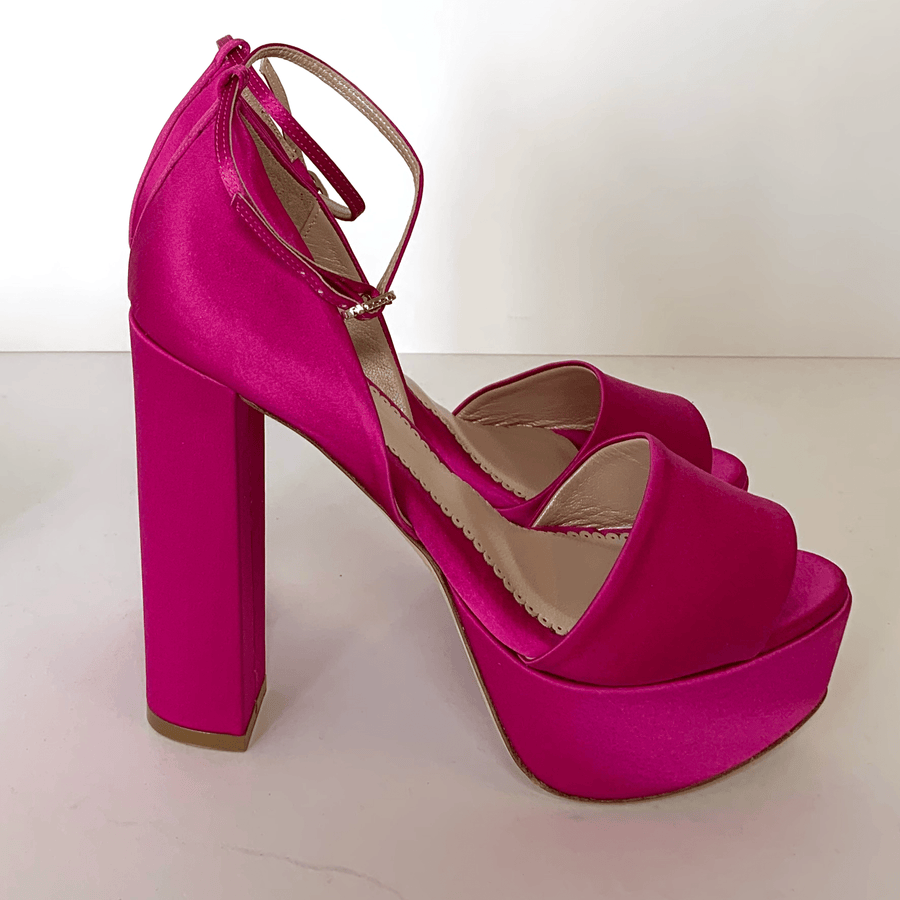 Fuchsia satin platform heels with a chunky block heel, featuring an ankle strap. The shoes are positioned on a white surface against a plain background, showcasing their vibrant color and sleek design. image
