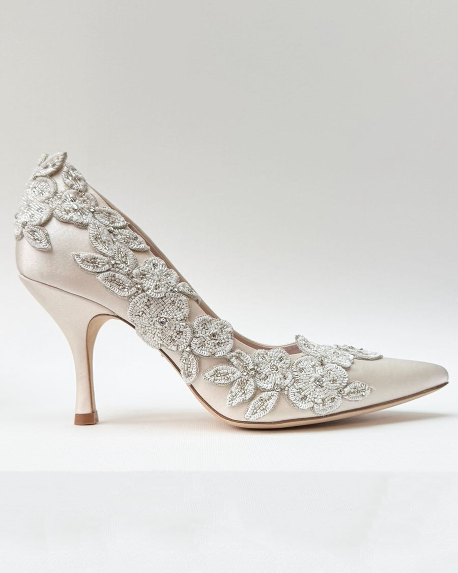 Elegant pointed-toe heels featuring intricate floral embellishments in a soft beige satin material. The shoes are set against a neutral background, showcasing their delicate design. The heel height is approximately 4 inches, providing a sophisticated lift. image