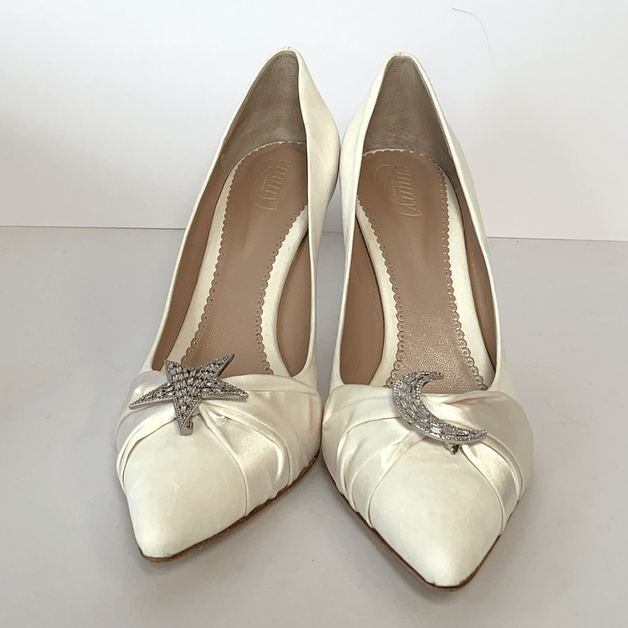 Elegant white heels featuring a pointed toe design, adorned with a star and crescent moon embellishment at the front. The shoes are displayed against a plain white background, showcasing their sleek silhouette. The angle captures the shoes from the front, highlighting the intricate detailing and smooth finish. image