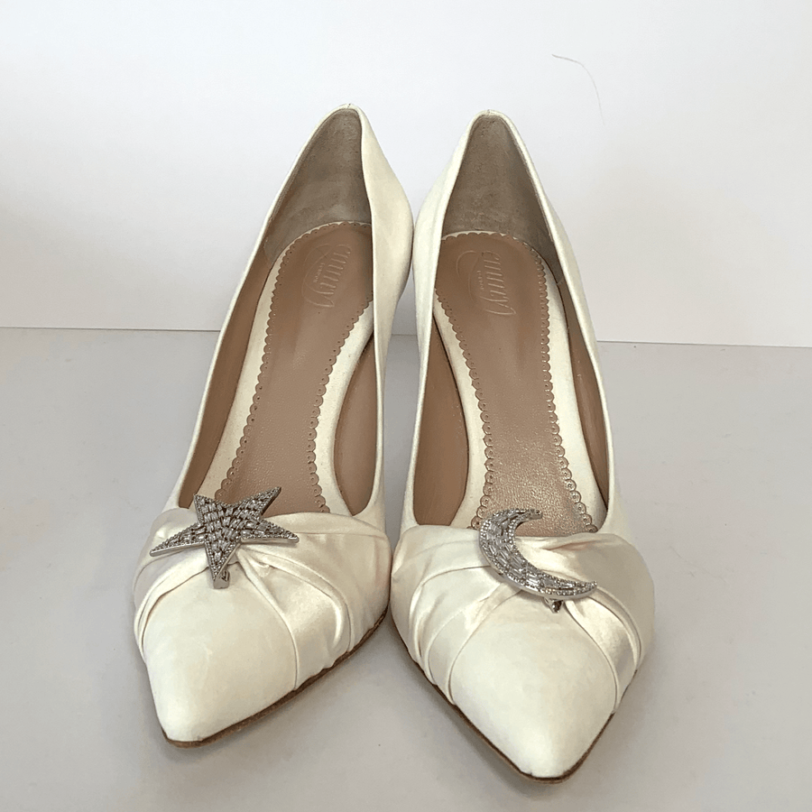 White heels with a pointed toe, featuring decorative silver star and crescent moon embellishments on the outer sides. The shoes are displayed on a light gray background, showcasing their elegant design. The angle captures both shoes side by side, highlighting the intricate detailing and smooth finish. image