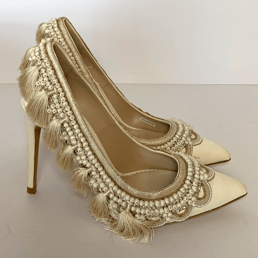 Elegant cream heels adorned with intricate beading and fringe detailing. The shoes feature a pointed toe and a high stiletto heel. The image is taken from a side angle against a plain white background, showcasing the luxurious embellishments. image