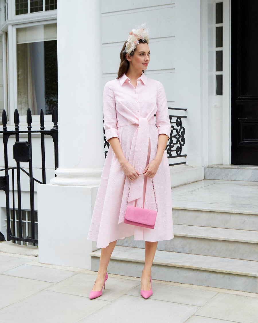 A woman stands elegantly on a marble staircase, wearing a light pink dress with a tied waist and three-quarter sleeves. She accessorizes with a matching pink handbag and a floral headpiece. The background features a classic white building with ornate black railings. The heels are a vibrant pink, complementing her outfit. image