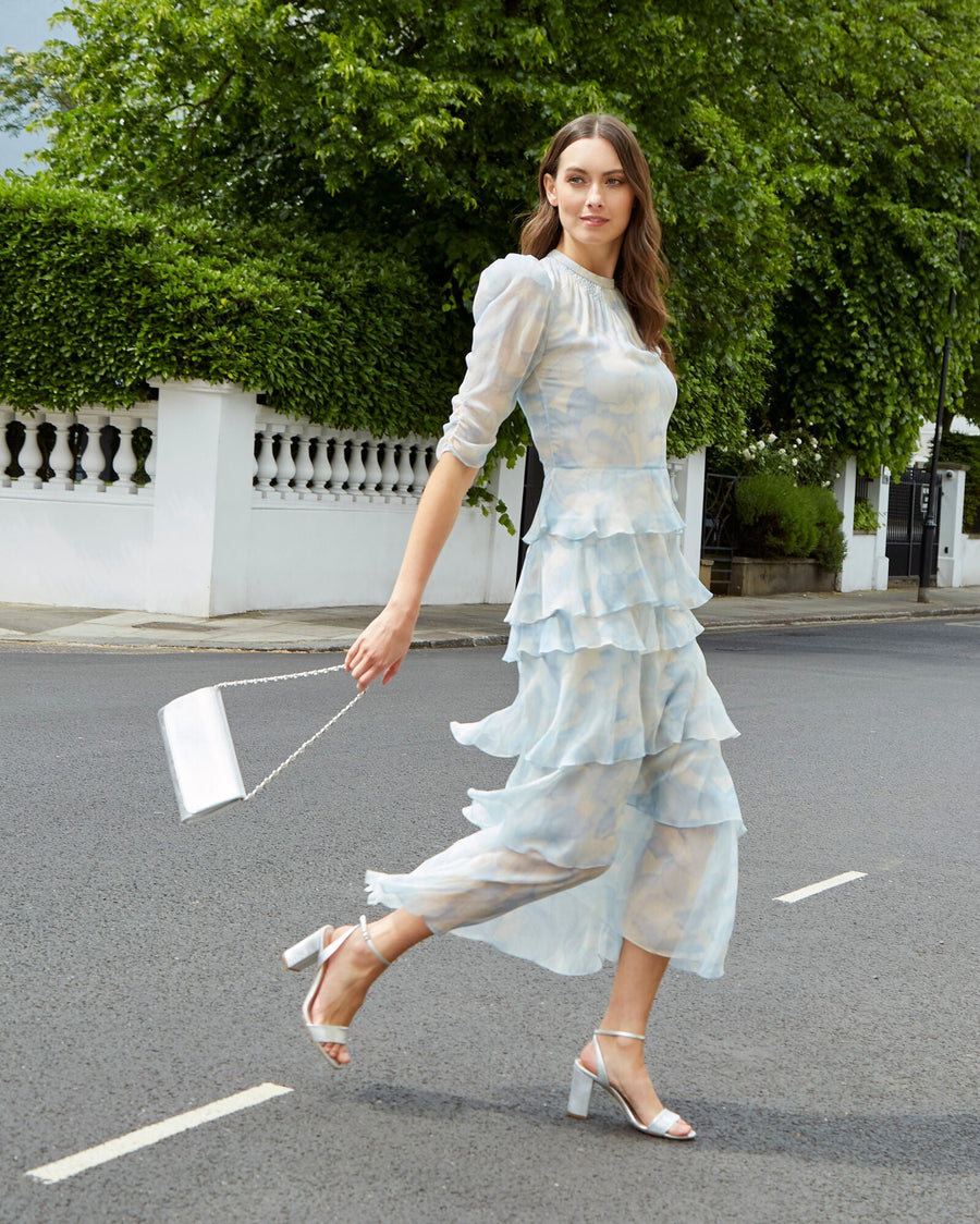 A model is walking on a street, wearing a light blue, tiered dress with ruffles. She is holding a silver clutch bag and wearing strappy heels. The background features greenery and a white wall, with a clear sky above. The heels are silver and have a moderate height. image
