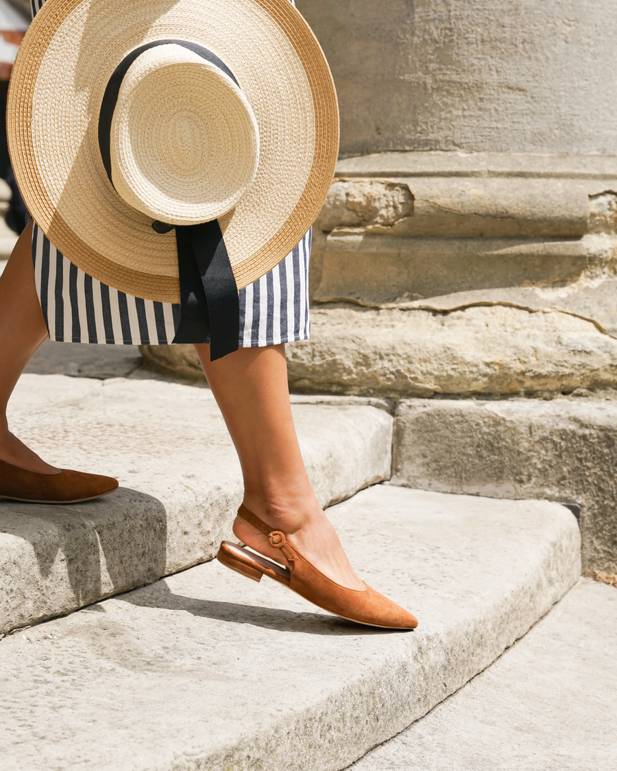 A woman is walking down a stone staircase, wearing a pair of brown suede slingback shoes with a pointed toe and a small heel. She is dressed in a striped skirt and is holding a wide-brimmed straw hat adorned with a black ribbon. The image captures her from a side angle, highlighting the shoes and the textured stone background. image