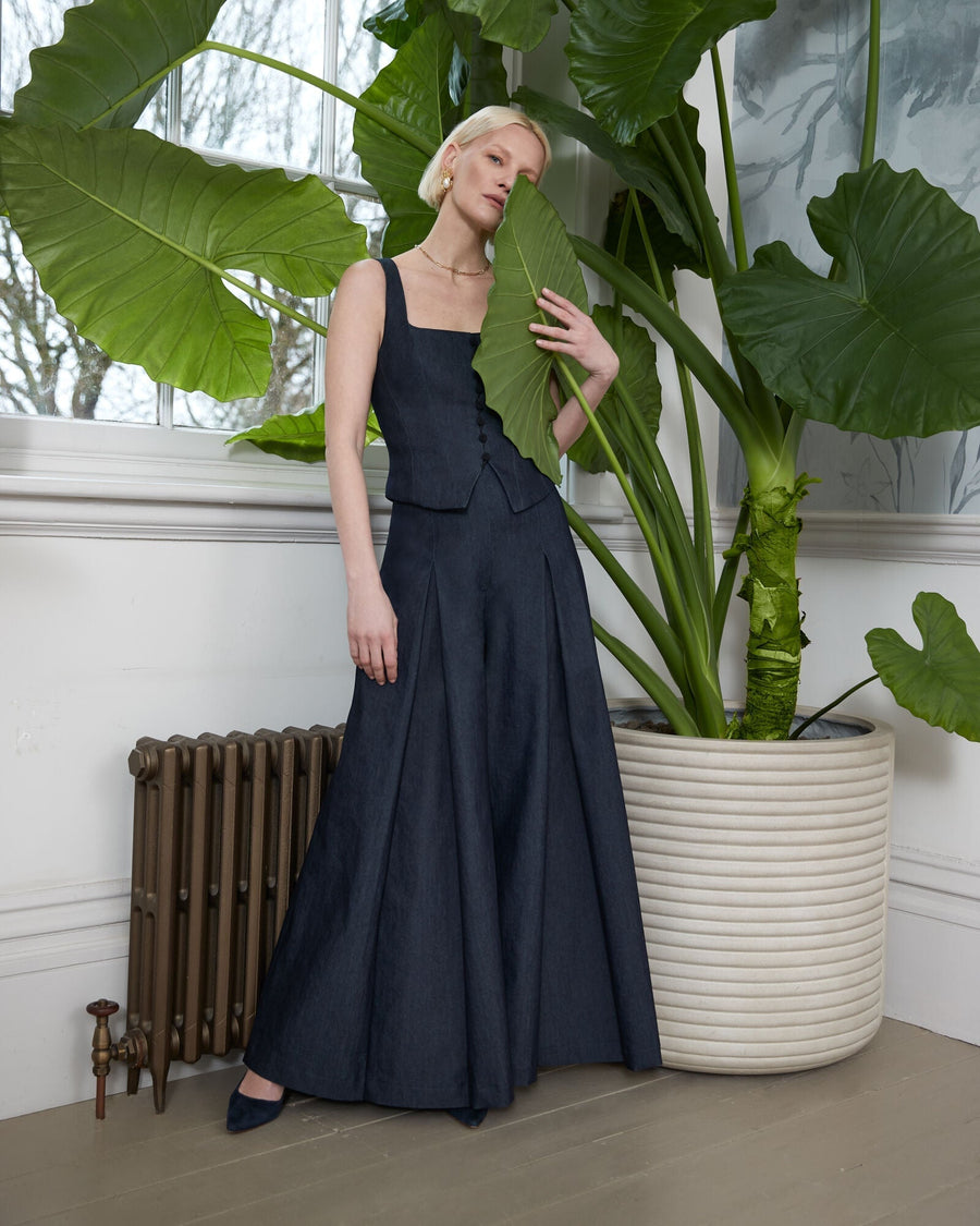 A model stands elegantly beside a large potted plant, wearing a fitted dark denim sleeveless top paired with a flowing, floor-length dark denim skirt. The outfit features a structured silhouette, accentuating the model's figure. The background includes a large window that allows natural light to illuminate the scene, highlighting the textures of the fabric. The model is wearing dark heels, completing the sophisticated look. image