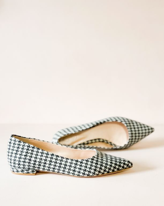 Pointed-toe flats made of tweed in a black and white houndstooth pattern. The shoes are positioned on a light-colored surface with a soft, neutral background. The angle showcases the shoes from the side, highlighting their low heel design. image