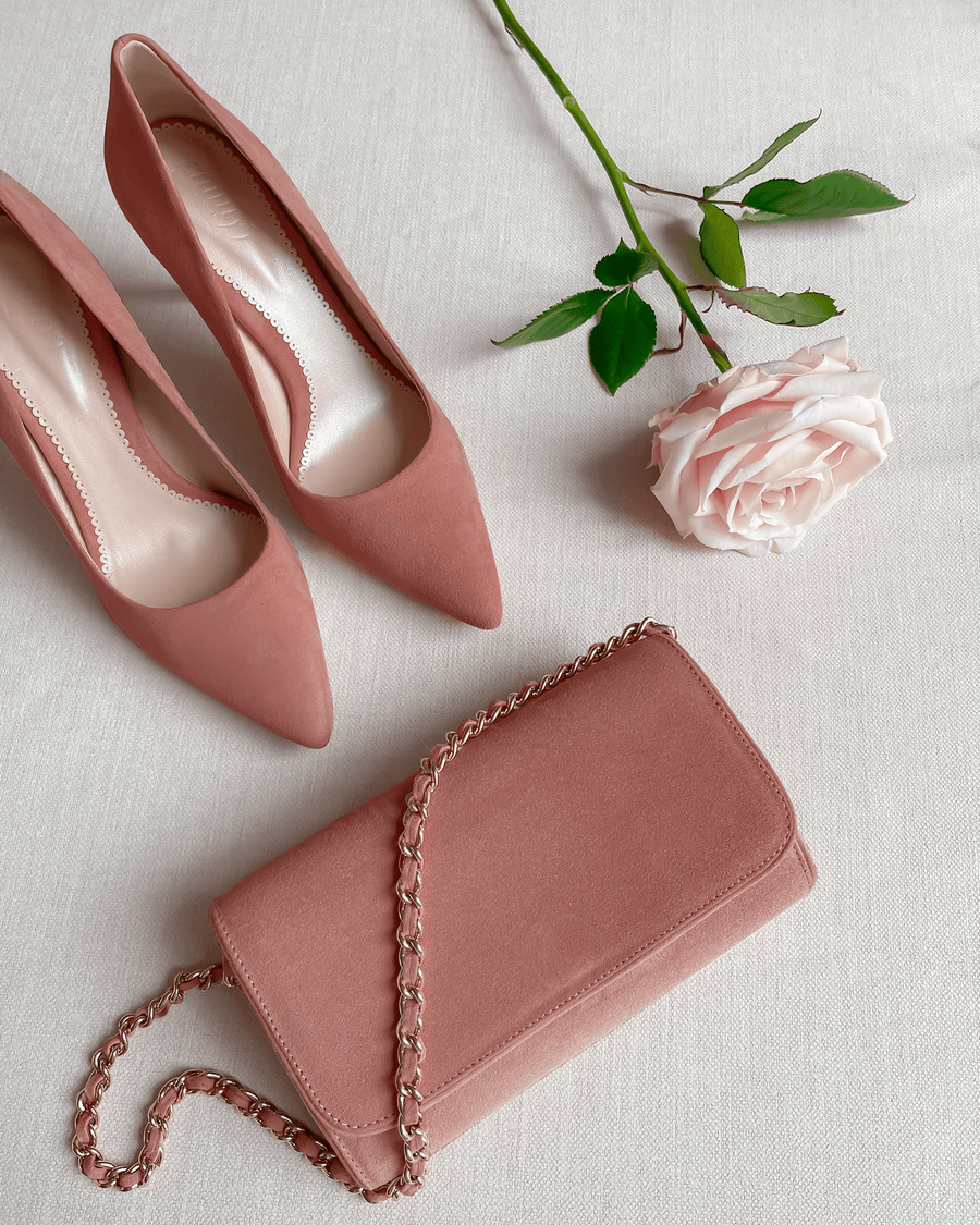 Pink suede heels with a pointed toe are positioned next to a matching pink clutch bag on a light-colored fabric surface. A single pink rose is placed nearby, adding a touch of elegance to the arrangement. The image is taken from a top-down angle, showcasing the shoes and accessories clearly. image