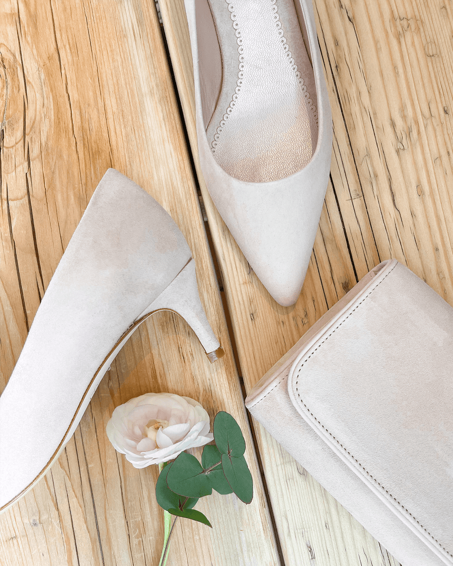 Elegant light beige suede heels with a mid-height heel, styled alongside a matching clutch and a delicate flower on a wooden surface. The image captures a top-down view, showcasing the pointed toe design and soft texture of the shoes. image