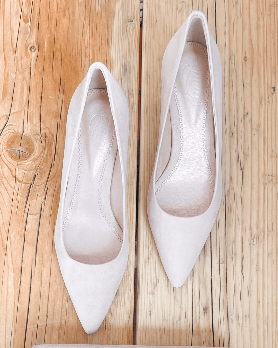 White suede pointed-toe heels displayed on a wooden surface. The shoes are shown from a top-down angle, highlighting their sleek design and smooth finish. The background features a light wooden texture, enhancing the minimalist aesthetic. image