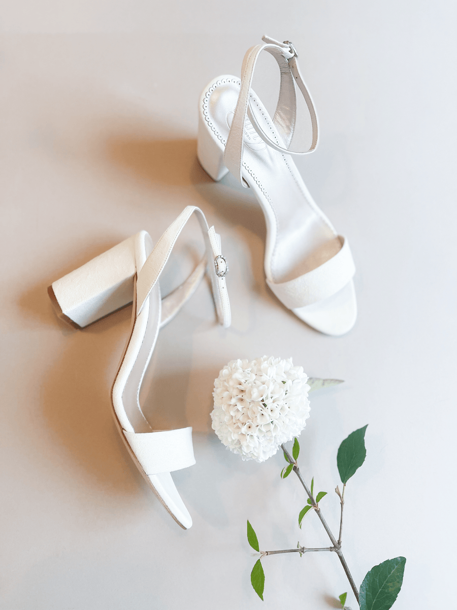 White block-heeled sandals with a wide strap across the toe and an adjustable ankle strap. The heels are mid-height and made of suede. The shoes are positioned on a light gray background, accompanied by a white flower and green leaves, creating a fresh and elegant aesthetic. image