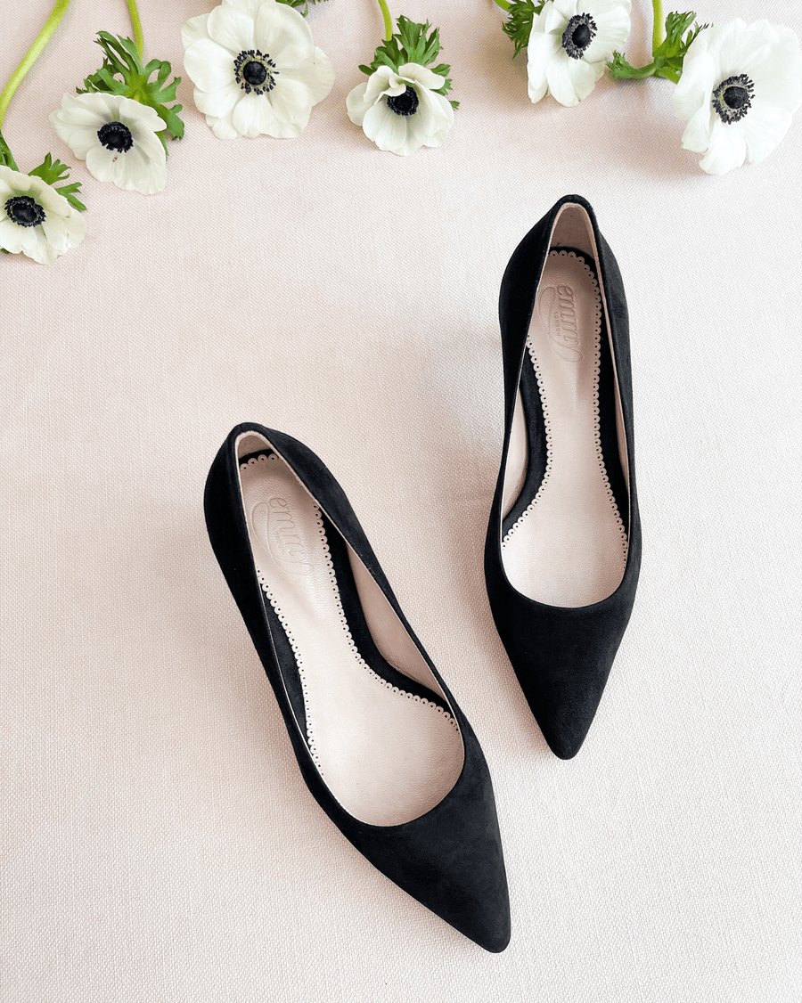 Black suede pointed-toe heels are displayed on a soft beige surface, surrounded by white anemone flowers. The shoes feature a sleek design with a low-cut vamp, showcasing their elegant silhouette. The image is taken from a top-down angle, emphasizing the shoes' refined shape and texture. image