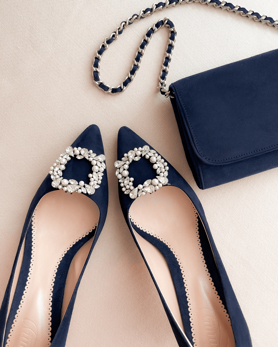 Navy suede pointed-toe heels featuring a decorative crystal buckle on the front. The shoes are positioned on a soft, neutral background, with a matching navy clutch purse placed beside them. The image is taken from a top-down angle, showcasing the elegant design and detailing of the shoes. image