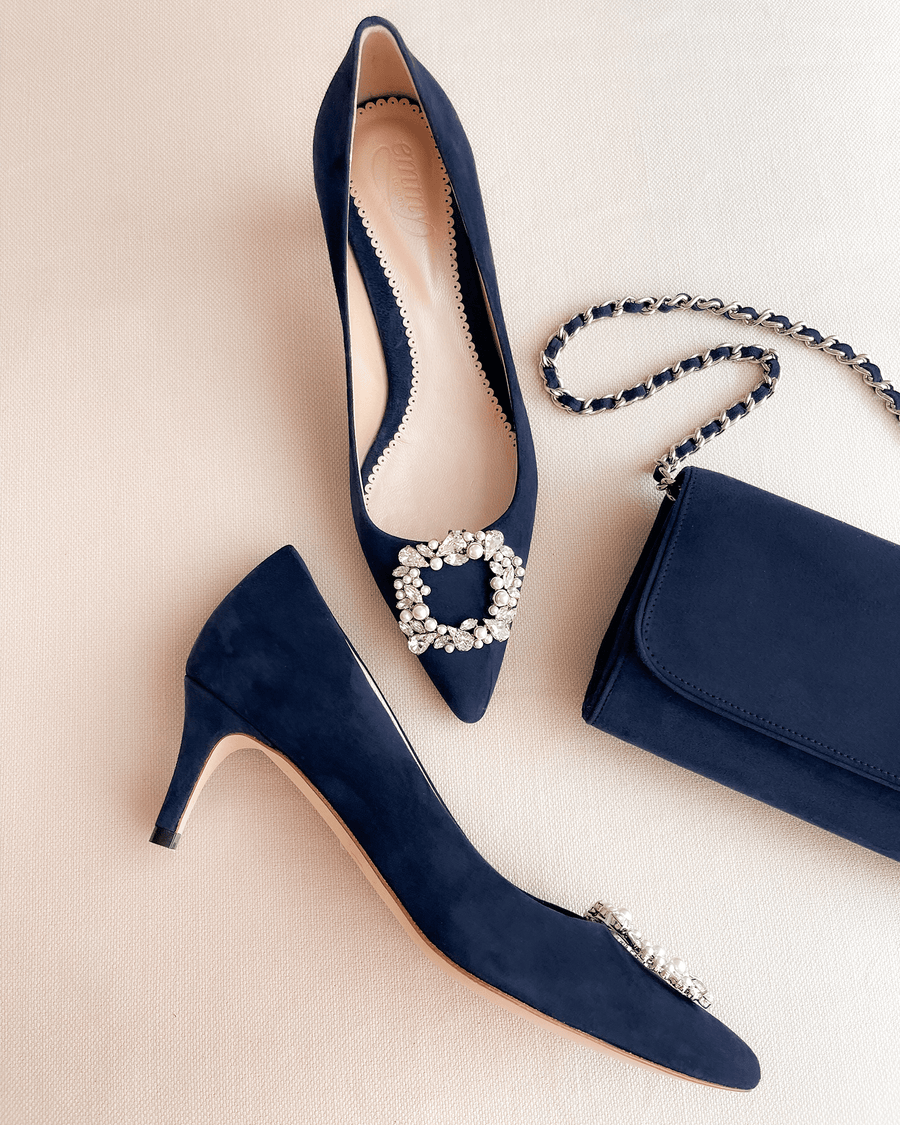 Navy blue suede heels with a pointed toe and embellished buckle detail, displayed alongside a matching clutch bag. The shoes are positioned on a light beige background, showcasing their elegant design. The heel height appears to be mid, complementing the overall sophisticated look. image