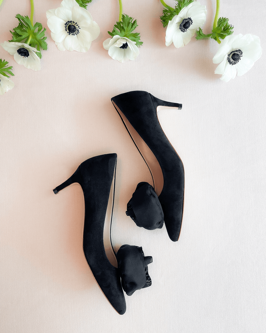 Black suede high heels are elegantly displayed on a soft, neutral background, surrounded by delicate white flowers. The shoes feature a pointed toe and a slender heel, showcasing a sophisticated design. image