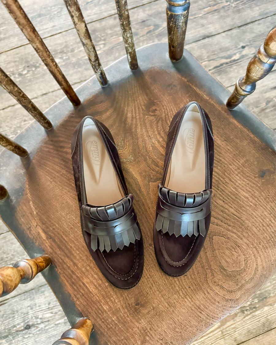 Dark brown suede loafers with a fringed leather detail on the front, positioned on a rustic wooden chair. The image is taken from a top-down angle, showcasing the shoes against a textured wooden floor background. image