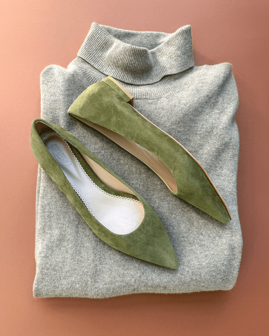 Green suede flats are displayed on a light gray sweater, showcasing a pointed toe design. The shoes are positioned at an angle, highlighting their sleek silhouette. The background is a soft, warm peach color, enhancing the overall aesthetic. image