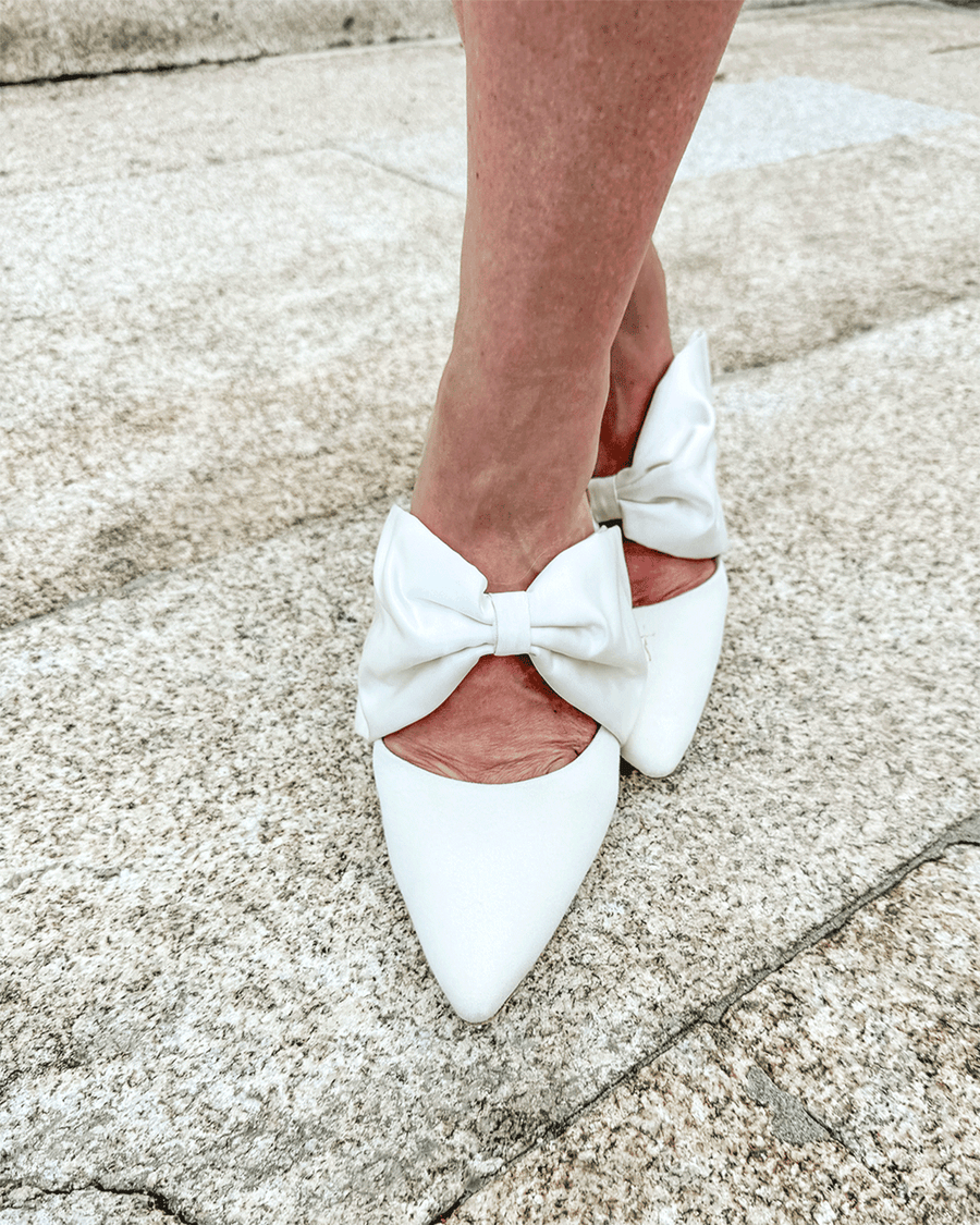 White pointed-toe heels with a large bow detail at the back, set against a textured stone background. The shoes are being worn by a person standing on a flat surface, showcasing the elegant design and clean lines of the footwear. image