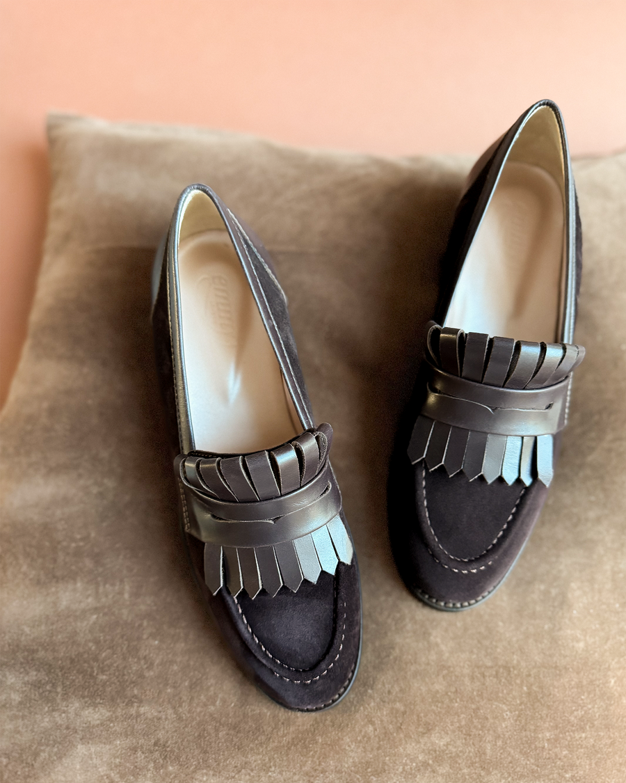 Stylish dark suede loafers featuring a decorative fringe detail on the front. The shoes are positioned on a soft, neutral-toned surface, showcasing their elegant design from a top-down angle. image