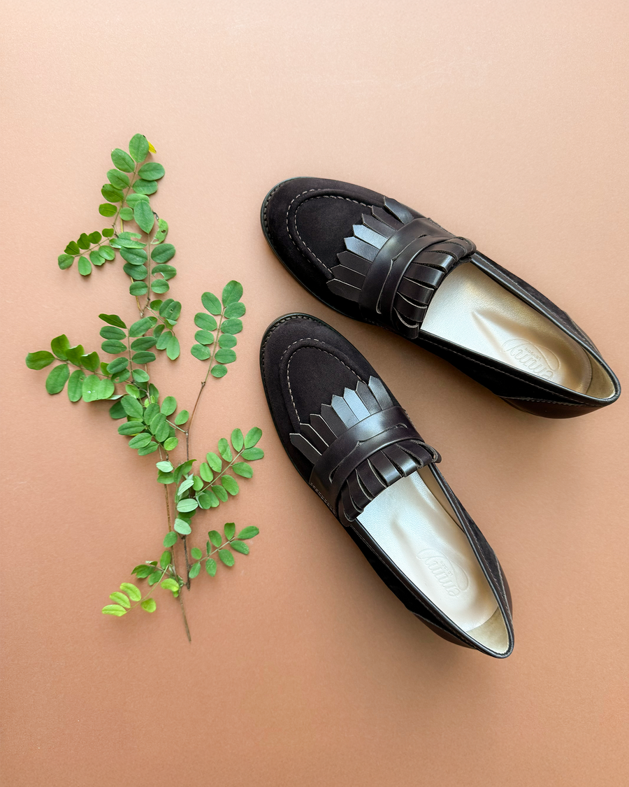 Black suede loafers with a decorative fringe detail, positioned on a warm brown background alongside a sprig of greenery. The shoes are displayed from a top-down angle, showcasing their sleek design and soft interior. image