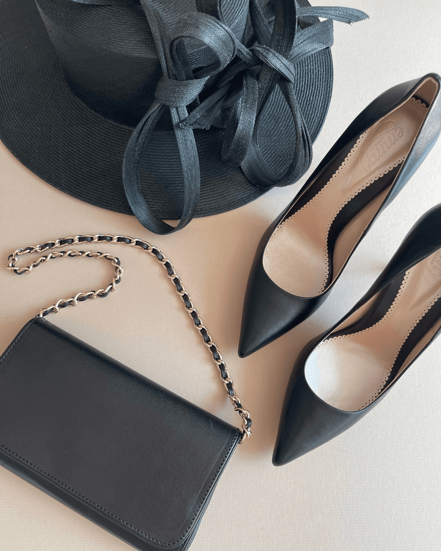 Black leather high heels with a pointed toe, placed next to a black chain-strap handbag and a wide-brimmed black hat. The items are arranged on a light-colored surface, showcasing a stylish ensemble. The angle captures the heels from the side, emphasizing their sleek design. image