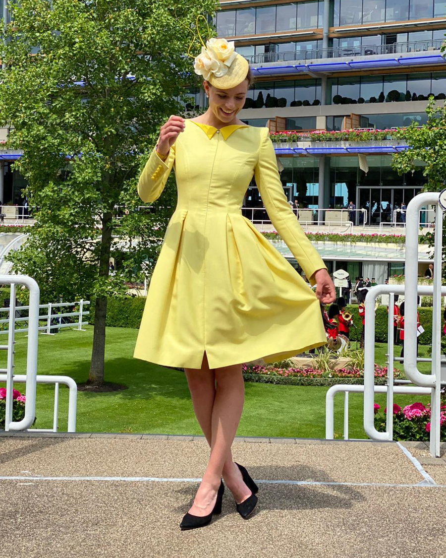 A woman is wearing a bright yellow dress with a fitted bodice and flared skirt, standing outdoors in a garden setting. She is smiling and playfully lifting the hem of her dress. The background features greenery and a modern building with people in formal attire. The shoes are black heels, complementing her outfit. image