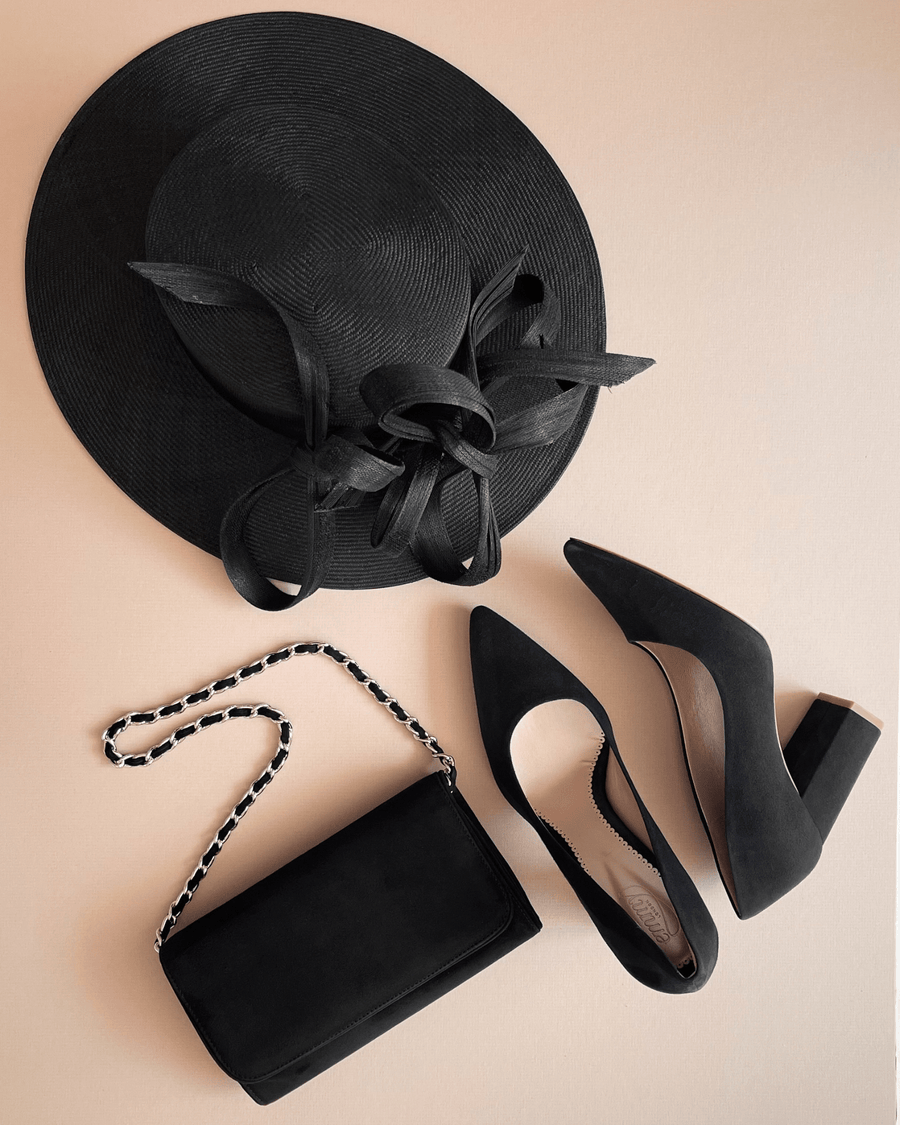Black suede heels with a block heel, approximately 3 inches in height, are positioned next to a black crossbody bag and a wide-brimmed black hat. The items are arranged on a soft beige background, showcasing a stylish ensemble. The angle captures the shoes from the side, highlighting their pointed toe design. image