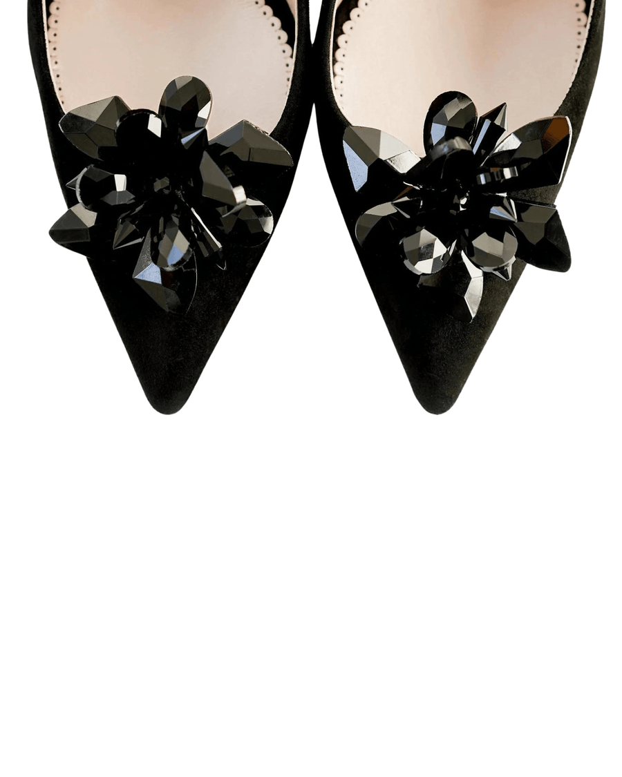Black suede heels with a pointed toe design, featuring a decorative black floral embellishment made of shiny materials. The image is taken from a top-down angle, showcasing the intricate details of the embellishment against a plain white background. image