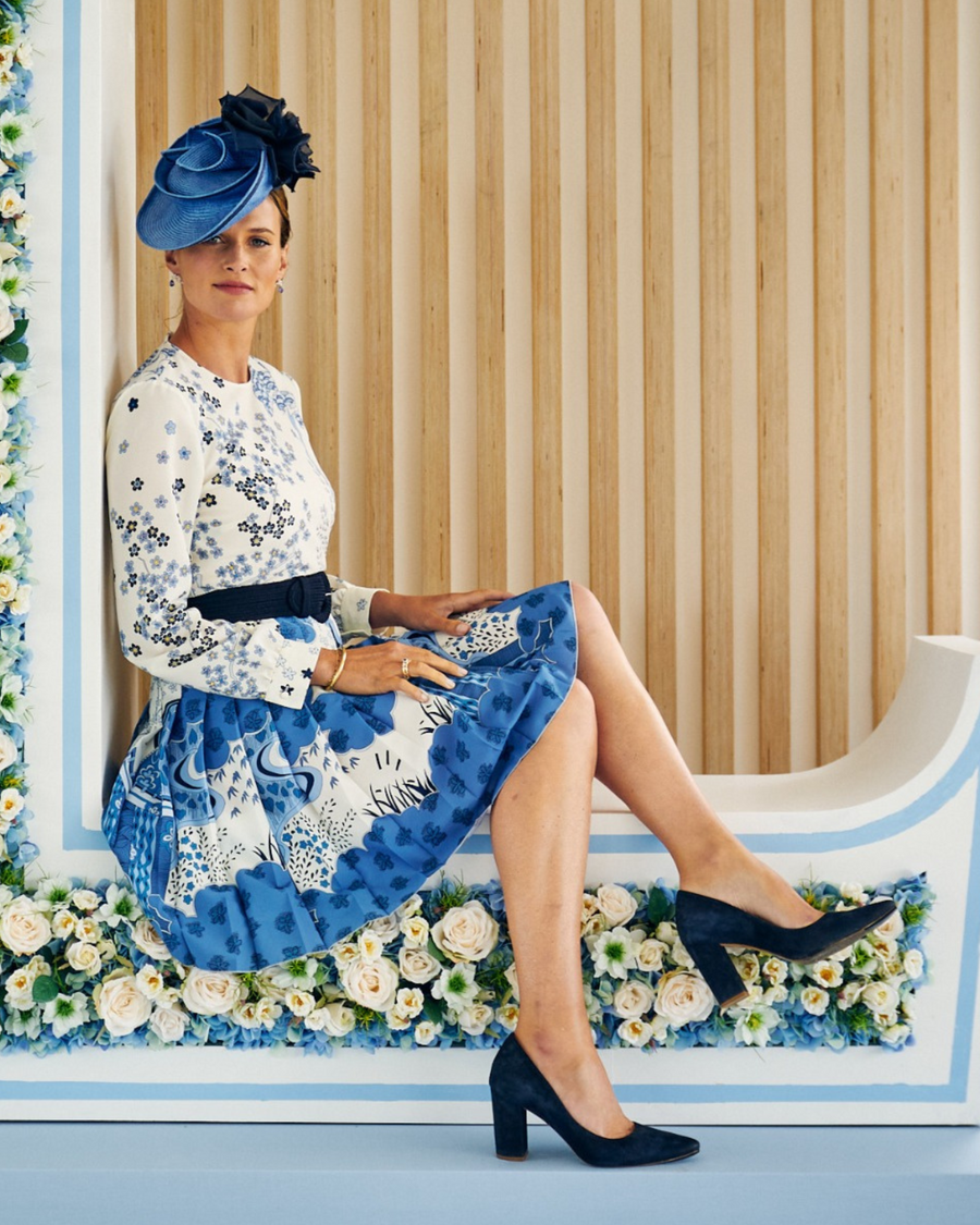 A woman is seated on a white bench adorned with flowers, wearing a floral blue and white dress paired with a matching blue fascinator. She is showcasing a pair of blue suede heels with a block heel design, positioned at an angle that highlights the shoes. The background features a wooden wall with floral decorations, creating an elegant atmosphere. image