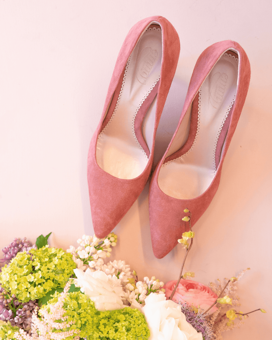 Pink suede high heels are displayed on a light background, surrounded by a bouquet of colorful flowers. The shoes feature a pointed toe and a sleek design, showcasing their elegant silhouette. image