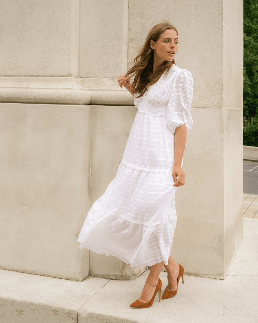 A woman is wearing a flowing white maxi dress with a tiered design, standing against a light-colored stone wall. She has long brown hair and is posing with one hand on her hip, showcasing a pair of high-heeled shoes made of suede in a warm brown tone. The background features greenery and a hint of a street, captured from a slightly angled perspective. image