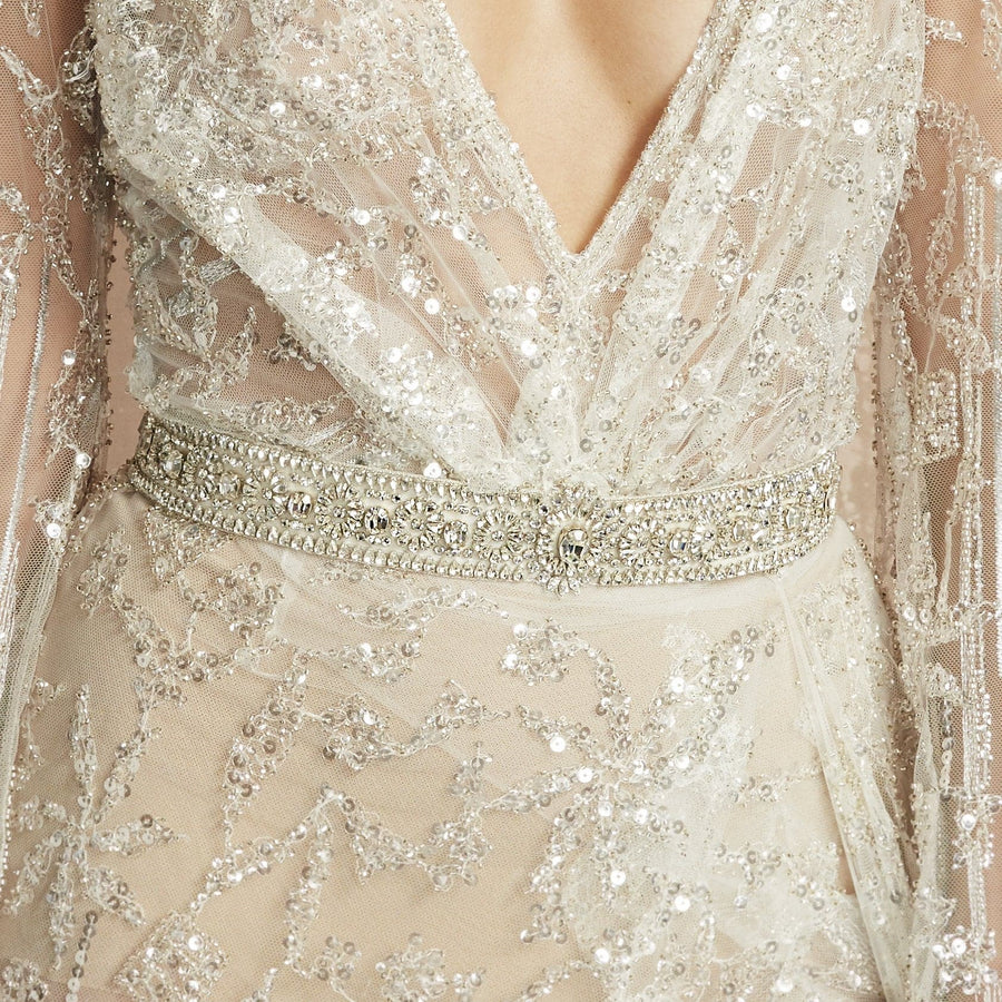The image features a close-up of a person wearing a stunning, intricately beaded dress with a deep V-neckline. The dress is adorned with delicate sequins and floral patterns, creating a shimmering effect. A sparkling embellished belt cinches the waist, adding elegance to the overall look. The background is softly blurred, focusing attention on the dress details. No shoes or heel height are visible in this image. image