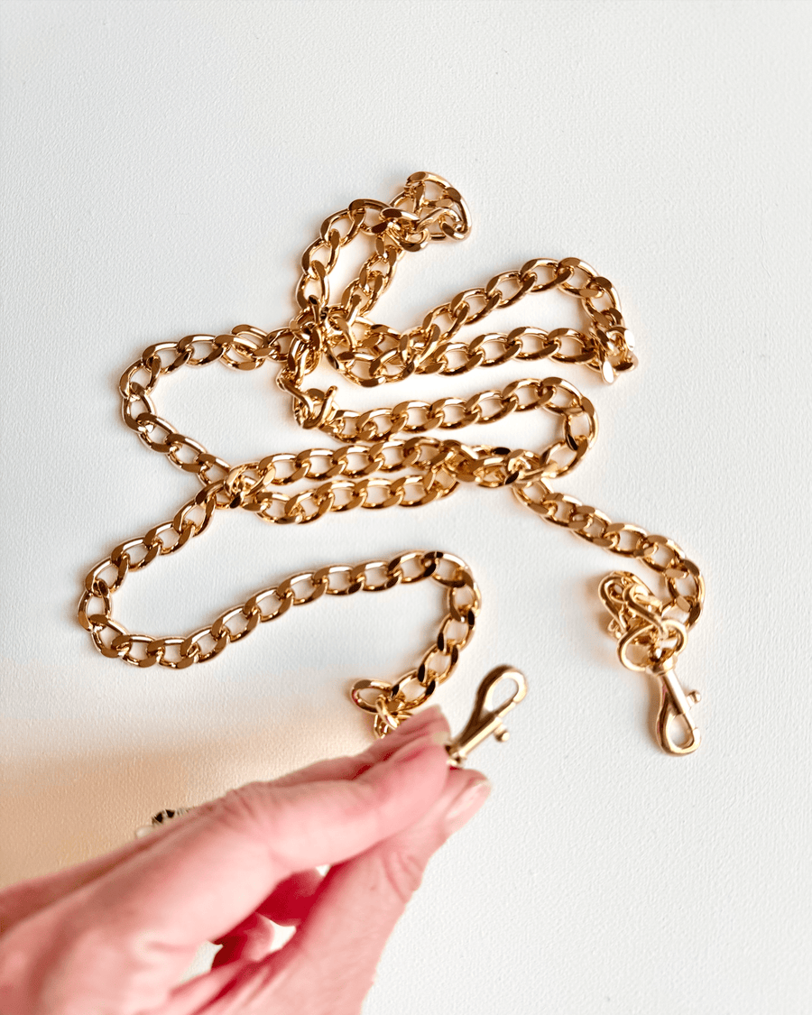 Gold chain with interlocking links laid out on a white background. A hand is holding one end of the chain, showcasing the clasp detail. The chain appears to be a decorative accessory, possibly for bags or jewelry. image
