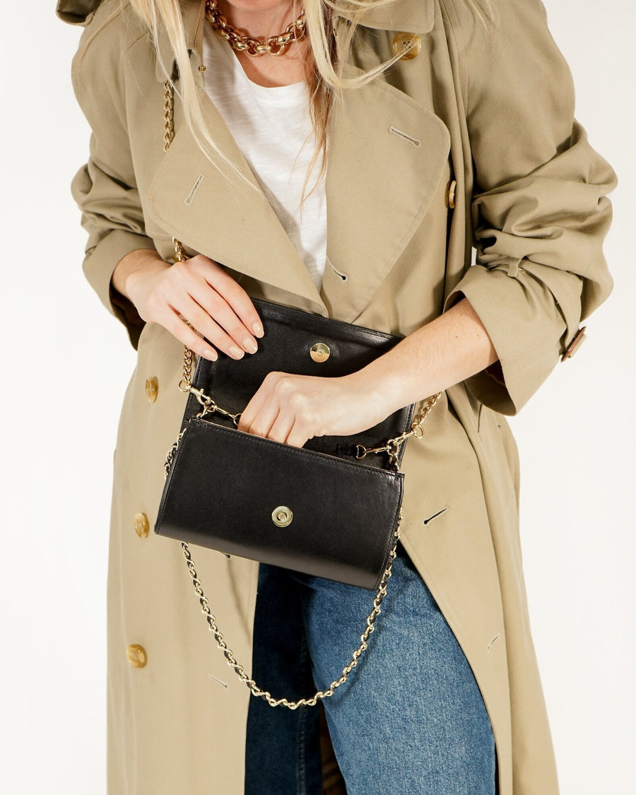 A model is wearing a stylish beige trench coat over a white t-shirt and blue jeans. She is holding a black handbag with a chain strap, which is partially open, revealing its interior. The background is a clean, neutral color, emphasizing the outfit and accessories. image