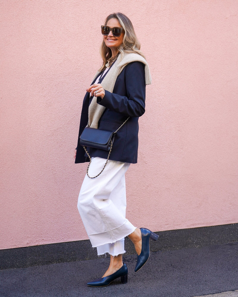 Elegant navy leather heels with a moderate block heel are worn by a woman standing against a soft pink wall. She is dressed in a stylish outfit featuring a tailored blazer, a light sweater draped over her shoulders, and white cropped pants. The angle captures her from the side, showcasing the shoes and the overall chic look. A small black crossbody bag completes her ensemble. image
