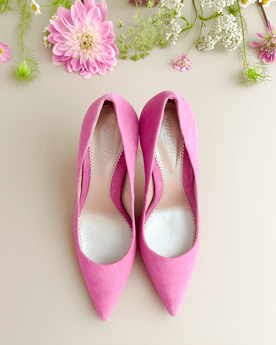 Pink suede heels with a pointed toe design are displayed from a top-down angle against a soft beige background. Surrounding the shoes are delicate flowers, including pink daisies and white blooms, adding a fresh and vibrant touch to the scene. image