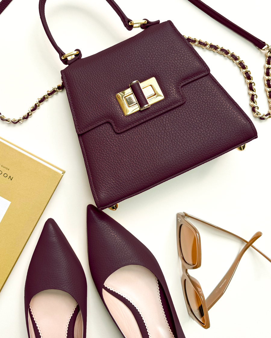 Elegant burgundy heels with a pointed toe design are positioned alongside a matching handbag, featuring a gold clasp, on a clean white background. The heels have a moderate height, complementing the sophisticated look. A pair of stylish sunglasses is also included in the arrangement, adding a chic touch to the overall display. image