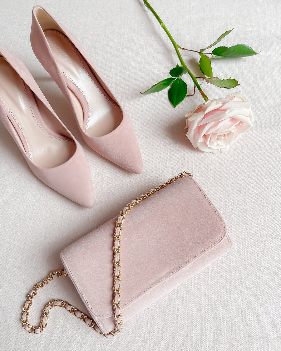 Elegant blush suede heels with a pointed toe design are positioned on a soft, neutral fabric background. A delicate pink rose and a small, chic clutch bag with a chain strap are artfully arranged beside the shoes, enhancing the overall sophisticated aesthetic of the image. image