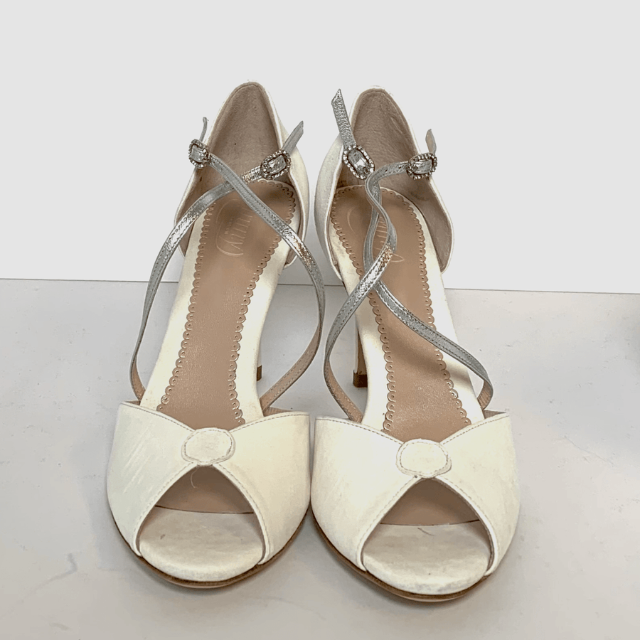 White open-toe heels with a delicate silver strap across the ankle. The shoes feature a rounded cutout design at the front and are positioned on a light background. The angle showcases the shoes from the front, highlighting their elegant style. The heel height appears to be mid. image