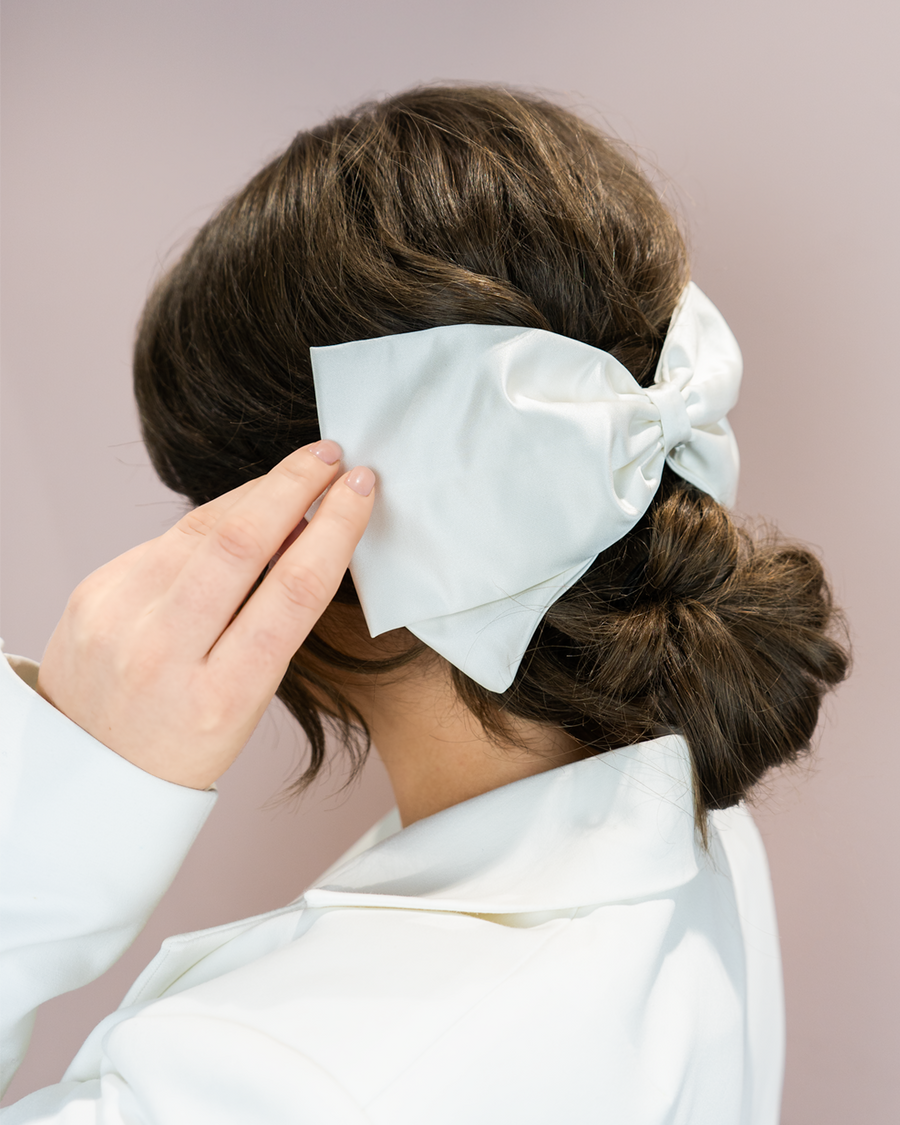 A woman with dark hair styled in a low bun is holding a large, elegant satin bow in a light shade of blue. She is wearing a white blazer, and the background is a soft, muted pink, creating a sophisticated and modern look. The angle of the image focuses on her hairstyle and the bow, highlighting the delicate texture of the fabric. image