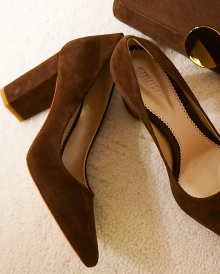 Brown suede high heels with a block heel, positioned on a soft, textured surface. The shoes feature a pointed toe and a sleek design, complemented by a matching brown handbag in the background. The image is taken from a top-down angle, showcasing the shoes prominently. image