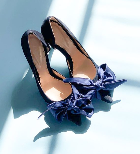 Elegant navy blue heels featuring a large satin bow at the front. The shoes are positioned on a light blue background, showcasing their sleek design. The angle captures the shoes from above, emphasizing the bow detail and the smooth finish of the material. image