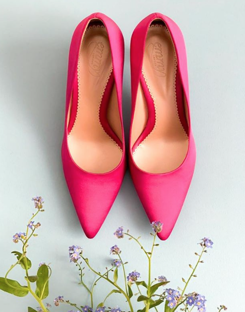 Pink pointed-toe heels are displayed on a light blue background, surrounded by delicate flowers. The shoes have a sleek design with a smooth finish, suggesting a mid heel height. image